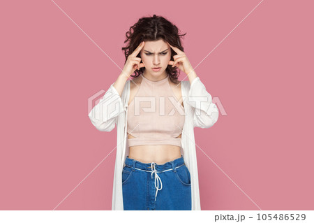 Portrait of confused puzzled woman with curly hair wearing casual style outfit thinking, keeps hands on temples, having serious expression. Indoor studio shot isolated on pink background. Portrait of confused puzzled woman with curly hair wearing casual style outfit thinking, keeps hands on temples, having serious expression. Indoor studio shot isolated on pink background. 105486529