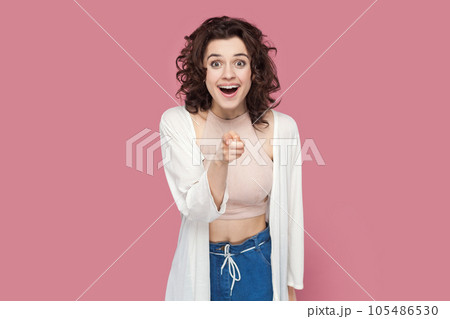 Portrait of amazed surprised woman with curly hairstyle wearing casual style outfit choosing you, looking at camera with big eyes and pleasant surprise. Indoor studio shot isolated on pink background. Portrait of amazed surprised woman with curly hairstyle wearing casual style outfit choosing you, looking at camera with big eyes and pleasant surprise. Indoor studio shot isolated on pink background. 105486530