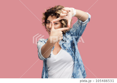 Portrait of concentrated attractive woman photographer with curly hairstyle wearing blue shirt standing looking at camera through frame of fingers. Indoor studio shot isolated on pink background. 105486531