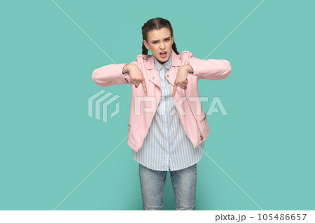 Portrait of strict bossy teenager girl with braids wearing pink jacket pointing both index fingers down, saying here and right now. Indoor studio shot isolated on green background. Portrait of strict bossy teenager girl with braids wearing pink jacket pointing both index fingers down, saying here and right now. Indoor studio shot isolated on green background. 105486657