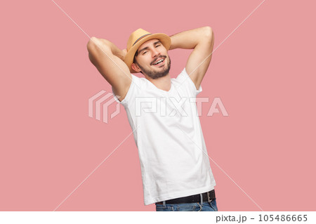 Portrait of satisfied relaxed handsome bearded hipster man in white T- shirt and casual hat standing with raised hands, looking at camera. Indoor studio shot isolated on pink background. 105486665