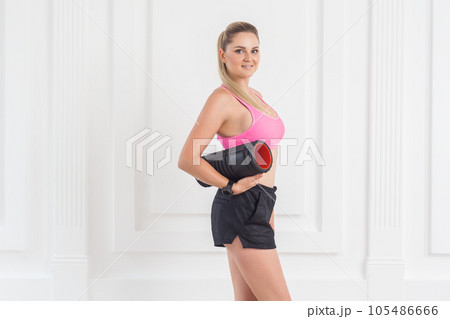 Portrait of athletic young beautiful blonde woman in pink top and black shorts standing, holding foam roll and looking at camera with toothy smile, enjoying her workout. Indoor studio shot. 105486666