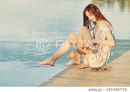 Young woman sits on edge of pier and takes off her sandals to soak her feet in water 105490739