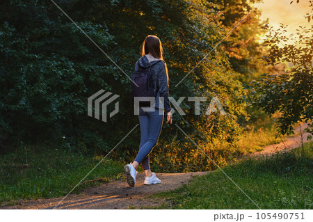 Back view of a woman goes along path from forest to clearing in rays of sunset 105490751