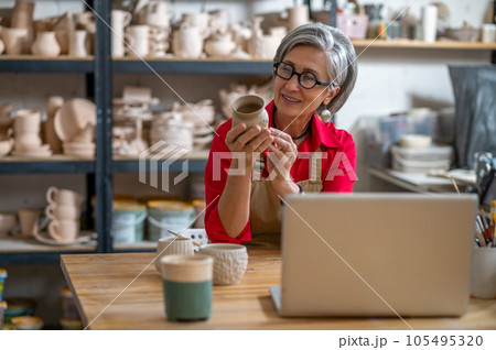 Mature woman painting pottery earthenware product in front of laptop while live streaming. Mature woman painting pottery earthenware product in front of laptop while live streaming. 105495320