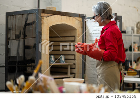 Mature craftswoman taking clay products from pottery kiln. 105495359