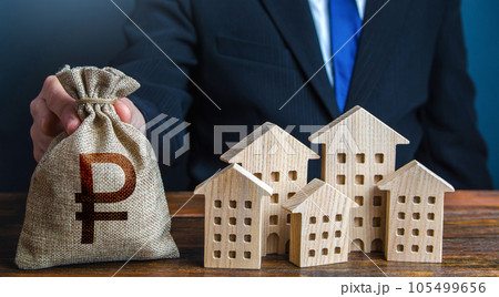 A businessman puts a russian ruble money bag near the houses. Investments in real estate assets. Municipal budget of the city. Official. Construction industry, rental business and hotel tourism. 105499656