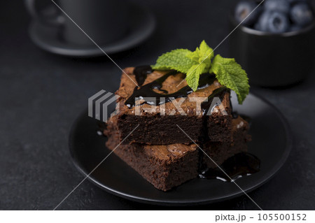 Chocolate brownie cake with mint, blueberries, chocolate topping and espresso. Traditional American dessert. Popular cake with dark chocolate on a black background. Close-up, selective focus. Chocolate brownie cake with mint, blueberries, chocolate topping and espresso. Traditional American dessert. Popular cake with dark chocolate on a black background. Close-up, selective focus. 105500152