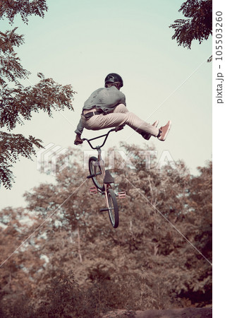 Back view dynamic image of young man in helmet riding bmx bike in forest, training, doing difficult and dangerous tricks Back view dynamic image of young man in helmet riding bmx bike in forest, training, doing difficult and dangerous tricks 105503260