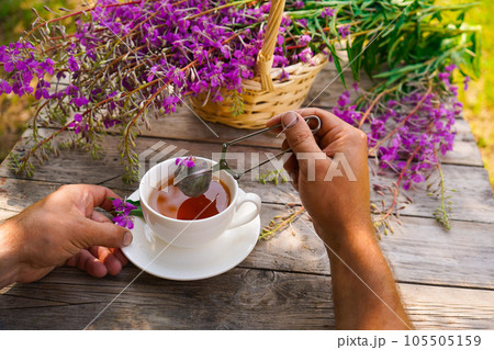 Close-up of men's hands with a white cup of tea. Healthy organic leaves Ivan chai tea Russian herbal on the wooden table Close-up of men's hands with a white cup of tea. Healthy organic leaves Ivan chai tea Russian herbal on the wooden table 105505159