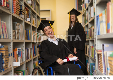 Happy young woman and woman in wheelchair in graduate gown with diploma in hands in library. Inclusive education. Happy young woman and woman in wheelchair in graduate gown with diploma in hands in library. Inclusive education. 105507670