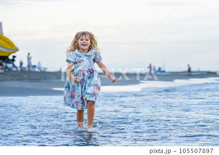 Happy Child, Little Preschool Girl in Dress Running And Jumping In The Waves During Summer Vacation On Exotic Tropical Beach by Sunset. Family Journey On Ocean Coast. 105507897