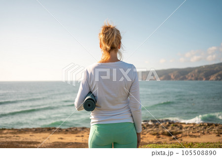 Senior woman holding yoga mat on ocean shore back view 105508890