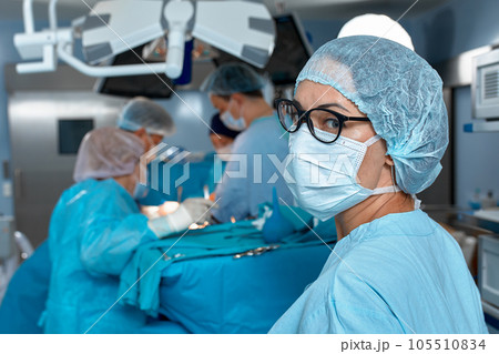 Close-up portrait of a female doctor in glasses and face mask on the background of surgeons team during complex surgical operation in a sterile operating room. Modern medicine, saving patients life. Close-up portrait of a female doctor in glasses and face mask on the background of surgeons team during complex surgical operation in a sterile operating room. Modern medicine, saving patients life. 105510834