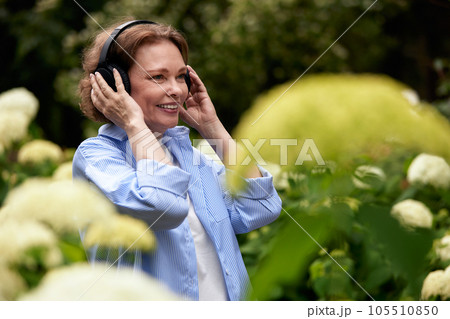 Portrait of attractive mature woman in summer park with wireless headphones. Senior lady enjoying music among flowers. Middle-aged brunette in blue shirt smiling happily. Active lifestyle for elderly. Portrait of attractive mature woman in summer park with wireless headphones. Senior lady enjoying music among flowers. Middle-aged brunette in blue shirt smiling happily. Active lifestyle for elderly. 105510850