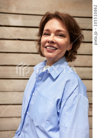 Closeup portrait of an attractive mature woman standing against background of siding cottage wall. Middle aged brunette female in a blue shirt smiling happily. Active lifestyle for the elderly. 105510862