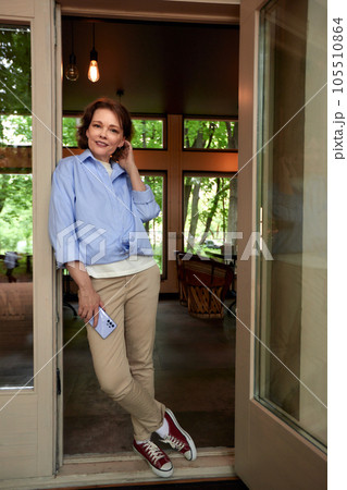 Mature woman in casual clothes on the porch of country cottage. Attractive middle-aged brunette with cell phone in her hand. Senior caucasian lady uses smartphone. Active lifestyle for the elderly. 105510864