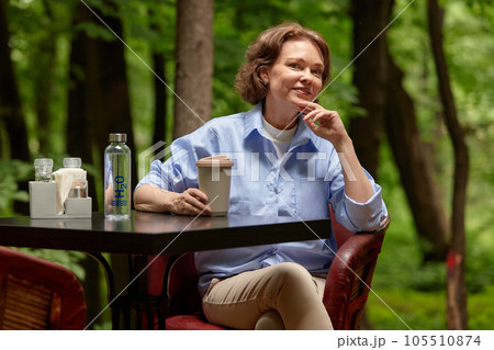 Adorable mature woman sits at the table in outside cafe with paper coffee cup and bottle of water. Senior lady enjoying life on summer terrace. Middle-aged brunette in blue shirt with dreamy smile. 105510874