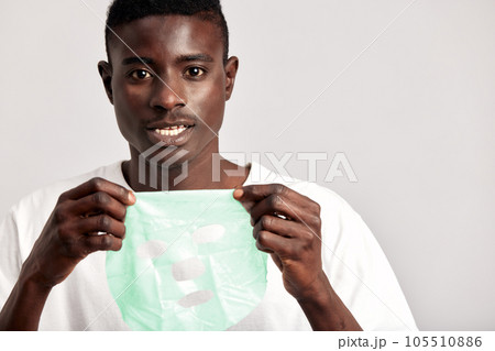 Studio portrait of young smiling black man applying moisturizing paper mask on his face. Happy African American millennial man uses beauty products in his skincare routine to maintain healthy look. 105510886