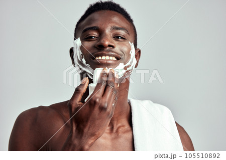 Studio portrait of handsome black guy with naked torso applying cream on his face. Afro American man with towel on his shoulder uses beauty products in his skincare routine to keep his healthy look. 105510892