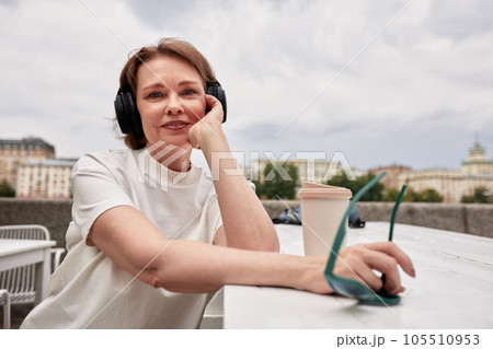 Adorable middle aged woman in white t-shirt, with sunglasses, wireless headphones and coffee paper cup sits at the table in city cafe. Mature woman enjoying life. Active lifestyle for the elderly. 105510953