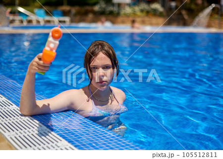 Young pretty caucasian girl with blue eyes shoots with a water gun in the pool. Pretty girl playing with water guns in pool. Cheerful children playing waterguns, rejoicing, jumping, swimming in pool. 105512811