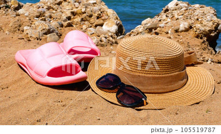 Pink beach clogs, straw hat and sunglasses on the sandy beach. In the background is the sea 105519787