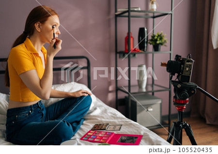Side view of beauty female blogger recording cosmetics product review, applying eyeshadow, filming video for channel using professional camera standing on tripod, sitting on bed in home studio. 105520435