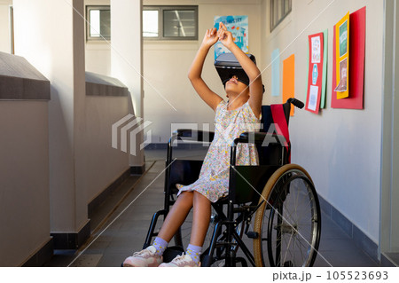 Happy biracial schoolgirl sitting in wheelchair and using vr headset at elementary school corridor Happy biracial schoolgirl sitting in wheelchair and using vr headset at elementary school corridor 105523693