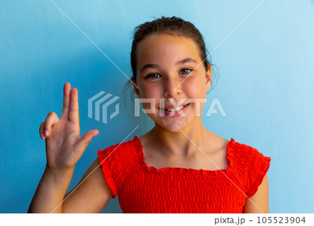 Portrait of happy caucasian schoolgirl doing sign language with hand over blue background 105523904