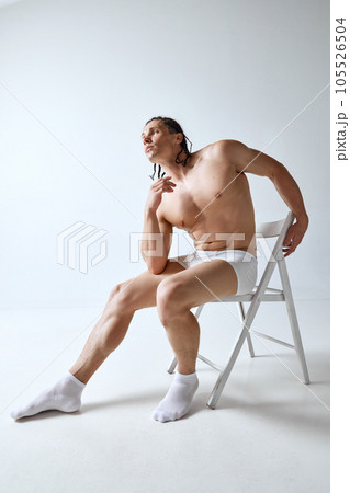 Handsome, stylish mature man posing shirtless in underwear, sitting on chair against grey studio background. Muscular body 105526504