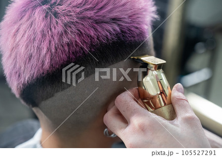 The hairdresser shaves the temple of a female client. Rear view of a woman with short pink hair in a barbershop. The hairdresser shaves the temple of a female client. Rear view of a woman with short pink hair in a barbershop. 105527291