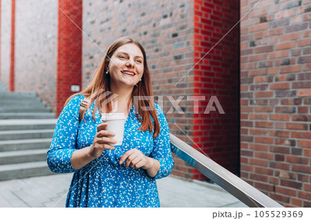 Cheerful young woman wearing blue dress walking outdoors, happy girl holding takeaway coffee cup on city background. Urban lifestyle concept. Cheerful young woman wearing blue dress walking outdoors, happy girl holding takeaway coffee cup on city background. Urban lifestyle concept. 105529369