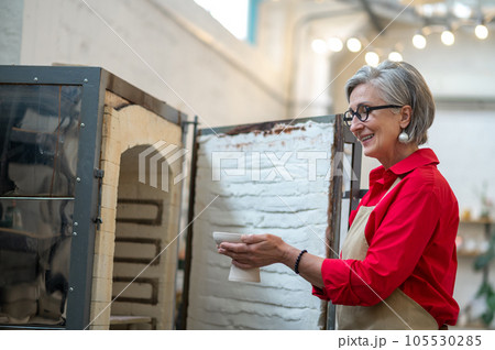 Positive craftswoman standing in front of open pottery kiln working in pottery workshop. 105530285