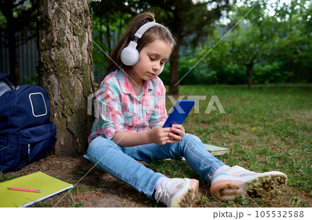 Cute little kid girl, primary school student holding a smart mobile phone, listening to music on headphones in the park Cute little kid girl, primary school student holding a smart mobile phone, listening to music on headphones in the park 105532588