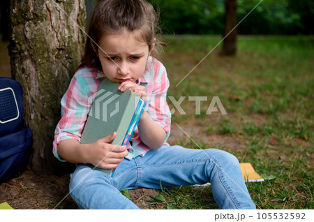 Adorable little kid girl holding textbooks, looking upset, sitting under tree in the backyard. Kids. Problems at school 105532592