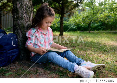 Little school girl reads a book in the park, sitting under tree with her backpack. Kids. Erudition. Education. Lifestyle 105532603