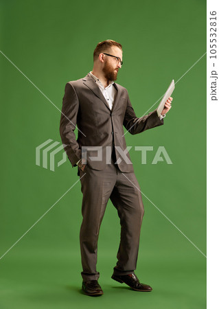 Full-length portrait of bearded businessman in suit and glasses seriously looking on tablet against green studio background Full-length portrait of bearded businessman in suit and glasses seriously looking on tablet against green studio background 105532816