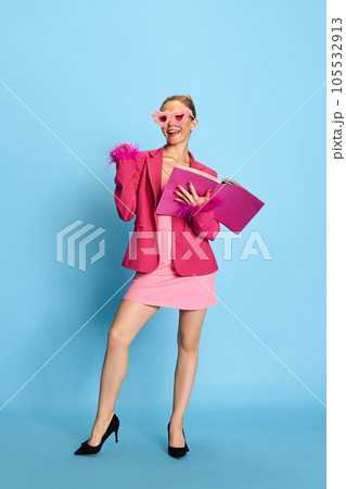 Photo of adorable girl, woman wearing glamor in unusual glasses, holding textbook smiling at camera on light blue studio background. Stylish young lady 105532913