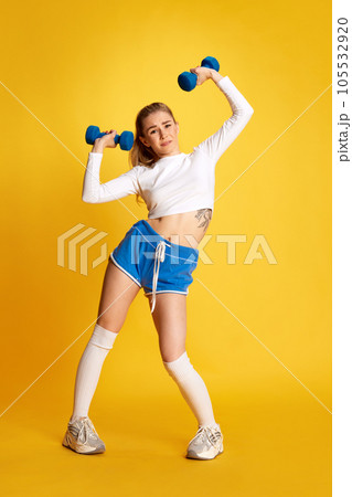 Full lenght of blonde girl trying to raise dumbbell looking at camera over bright yellow background. 105532920