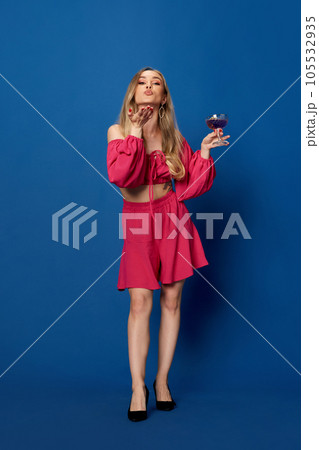 Full lenght of amazing woman in bright clothes holding cocktail and send air kiss at camera over blue studio background. 105532935