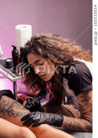 Female tattooist creating black and white flower design on thigh of client Female tattooist creating black and white flower design on thigh of client 105533054