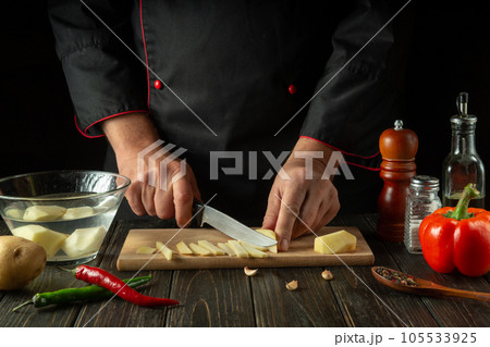 The cook cuts the potatoes into small pieces. Hands of a chef with a knife while preparing delicious food The cook cuts the potatoes into small pieces. Hands of a chef with a knife while preparing delicious food 105533925