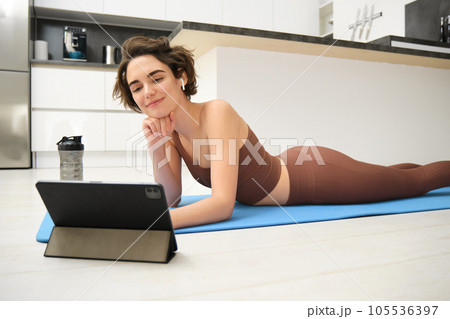 Portrait of smiling girl in fitness tracksuit, lying on mat at home, looks at tablet, watches online sports video tutorial, meditaiton lesson, yoga or workout online gym instructor Portrait of smiling girl in fitness tracksuit, lying on mat at home, looks at tablet, watches online sports video tutorial, meditaiton lesson, yoga or workout online gym instructor 105536397