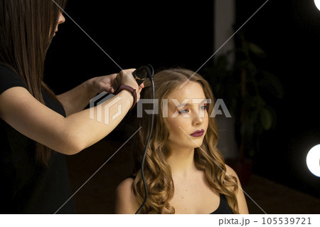 Professional stylist hairdresser curls long female hair with curling iron and makes styling for woman client. Beauty salon. Professional stylist hairdresser curls long female hair with curling iron and makes styling for woman client. Beauty salon. 105539721