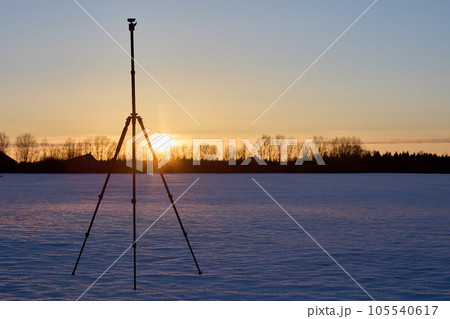Photographer equipment, an aluminum tripod for camera is set on snowy crust in field after sunset. 105540617