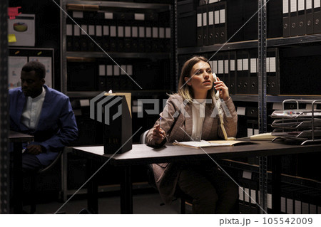 Manager having remote talk with businessman, working overtime at accountancy report arhive room. Confident repository employee organizing administrative files, searching for management research 105542009