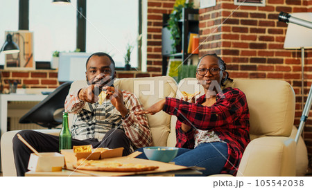Young couple eating slices of pizza and watching show on television, laughing and being together. Man an woman in relationship enjoying takeaway meal to watch favorite film on tv. Tripod shot. Young couple eating slices of pizza and watching show on television, laughing and being together. Man an woman in relationship enjoying takeaway meal to watch favorite film on tv. Tripod shot. 105542038