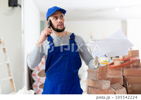 Master in blue overalls talking on mobile phone, discussing documents in renovated building Master in blue overalls talking on mobile phone, discussing documents in renovated building 105542224