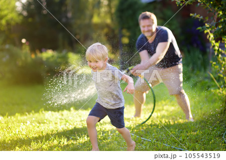 Funny little boy with his father playing with garden hose in sunny backyard. Preschooler child having fun with spray of water. 105543519